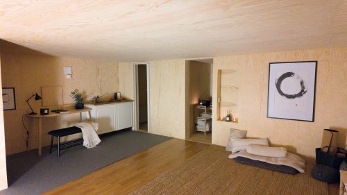 Bright wood-lined retreat room with a low bed, kitchenette and open doorway.