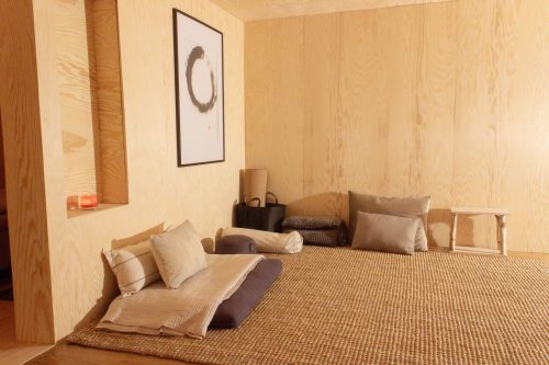 Low bed with beige cushions on a woven rug in a warm wood-lined retreat room.